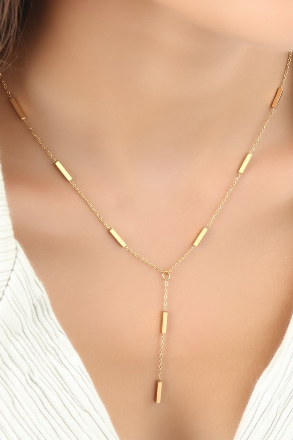 Model mit weißem, geripptem Oberteil trägt eine goldene Halskette mit rechteckigen Gliedern und Y-förmigem Anhänger. Das Bild zeigt eine Nahaufnahme des Halses und Dekolletés.